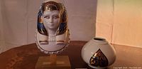 Porcelain bust of Egyptian Pharaoh with 24k gold and blue detailing on wooden base, next to round white vase with gold and blue decoration