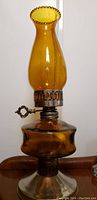 Full view of antique amber glass oil lamp showing tall amber chimney with scalloped top, amber glass reservoir, metal burner with wick control, and clear glass base with a metal bottom.