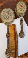 Vanity set showing mirror, brush, and comb with intricate bronze detail and floral fabric backs.