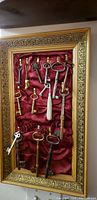 Full view of collection of 16 antique keys mounted on red fabric inside an ornate gold frame
