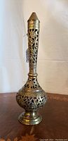 Front view of tall brass decorative piece showing intricate floral cut-out design on elongated neck and bulbous body.