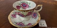 Photo showing antique Japanese porcelain teacup with pink and purple flowers, gold gilded rim and handle, sitting on matching saucer with similar floral design and gold trim.