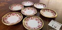 Seven small round porcelain dishes with red and pink floral patterns on the rim and gold trim, displayed on a wooden surface.