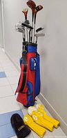 Front and side view of red and blue golf bag with multiple irons and woods inside, plus included yellow and black knitted club covers on floor.