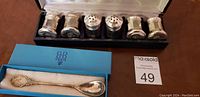 Front view of six silver shakers in original black and velvet-lined presentation box with silver company branding inside lid. Original blue velvet box with antique silver spoon in foreground.