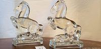Pair of clear crystal horse figurines on rectangular bases, shown from front angle against light background.