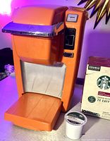 Orange Keurig coffee maker with closed lid and drip tray extended; Starbucks Dark Roast coffee pod box and one individual pod visible on table.