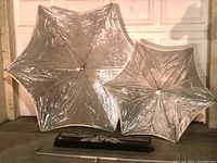 Two silver reflective umbrella reflectors hexagonal in shape, larger and smaller sizes, with packaging and a bracket on the floor beneath them.