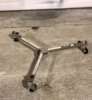 Photo of the unfolded SLIK Universal Tripod Dolly U700 on a concrete floor showing its three aluminum legs and caster wheels.