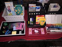 Overview of school and art supplies including glue sticks, notebooks, crayons, chalk, paint palette, and Hello Kitty pencil box organized in trays