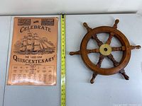 Copper Quincentenary celebratory sign and small wooden ship's wheel decor shown together on table.