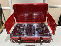 Open view showing two metal burners, control knobs, foldable side wind guards, and red enamel finish.