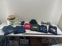 Overview of lot showing two women's hats, four embroidered T-shirts, two pairs of men's jeans, umbrella, gloves, reversible belt, wall rack and 'Style' sign on a table.
