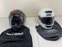 Two motorcycle helmets side by side on top of branded bags - left Storm with red stripes, right white Shoei full face helmet