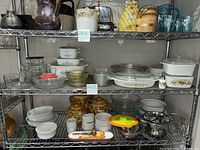 Shelf with assorted vintage glassware and kitchen items including white Corning Ware casserole dishes with blue floral pattern, clear glass bowls and casserole dishes, amber and green textured glass bowls, ramekins, and pie plates.