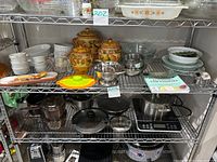 Shelving unit with the ceramic canisters with pear design, white Corelle dishes, metal bowls, silicone lids, wooden spoons, colander, and Pyrex measuring cups and bowls with the Date Night Cook Book.