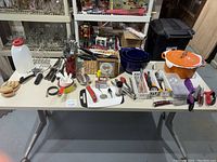 Full view of table with various kitchenware items including large orange stock pot, knives, cutlery, bowls, and utensils