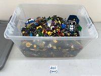 Large clear plastic container filled with assorted LEGO bricks, wheels, mini-figure parts, and small assembled portions.