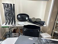 Photo showing disassembled components including collapsible frame, black oval case with halogen lights and cords, channel bars in bags, and carpet pieces.