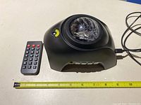 Black party laser strobe light with faceted dome lens and four buttons on front next to measuring tape for size reference, with remote control placed beside it.