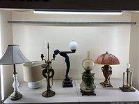 Seven lamps arranged left to right: crystal base lamp, ceramic lamp, brass harp frame lamp, art deco figure lamp, porcelain oil lamp, ornate vase-style lamp, candelabra style lamp