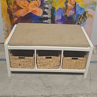 Front view of white wooden bench with beige removable cushion and three woven storage baskets below