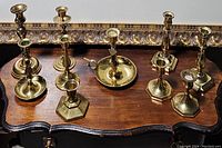Eight brass candle stick holders arranged on wooden surface with ornate frame in background