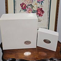 Two white Caring Collection branded boxes, one large and one small, on a wooden table with floral framed artwork in background.
