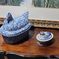 Photo showing both chicken dishes on a wooden surface with framed painting in background. Larger speckled dish on left, smaller detailed dish on right.