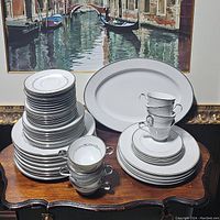 Two stacked sets of white china with silver rim accents including plates, cups, and serving platters on a wooden table.
