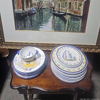 Photo showing multiple hand-painted plates and a small bowl stacked on a wooden table beneath a framed picture, highlighting colorful folk art designs.