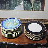 Photo shows two stacks of china plates. The left stack has a floral design with the word Rosemary, the right stack has black rim with gold trim.