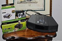 Lot contents on wooden surface against framed artwork: Bose Wave Radio CD, Sony Cyber-shot camera, VuPoint Photo Cube printer with box, and photo cartridge box.
