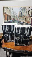 Two unopened boxes of Daniela brand six-piece goblet sets on a wooden table with a Venice backdrop.