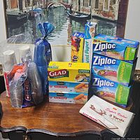 Wide view showing all items on table including plastic cups, storage bag boxes, parchment paper, and plastic wrap under framed artwork.