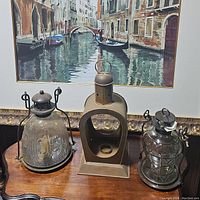 Three lantern candle holders displayed on a wooden surface in front of a framed painting.