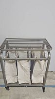 Front view of metal laundry cart with three off-white removable laundry bags and mesh top shelf.