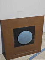 Front view of metal art piece showing rust-toned square base with centered black square and raised blue circular element.