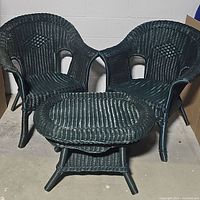 Two black wicker chairs with curved armrests and a small oval matching wicker table set together on concrete floor against white wall.