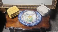 Top view showing two stacks of small square plates, one yellow set and one white set, alongside a large round serving plate with floral design