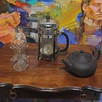 Full view of the set showing the glass decanter, French press, and black teapot on a wooden table with a vibrant painting in the background.