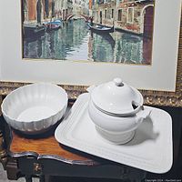 Photo showing white ceramic scalloped bowl, covered soup tureen with ladle on rectangular serving platter placed on wooden table.
