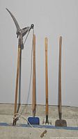 Five garden tools against wall: pruning saw, hoe, cultivator, shovel, and wooden tool with rope