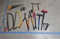 Photo showing various hand tools including saws, hammers, pruning shears, scrapers, and protective goggles arranged on the floor.