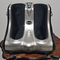 Front view of UComfy foot massager in silver plastic housing with black fabric foot chambers on wooden surface.