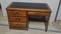 Front view of vintage wooden desk showing black top and four drawers with oval handles, right side has turned wood legs.