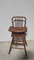 Front view of vintage wooden high chair showing the contoured seat, tray, spindled backrest, and safety strap.