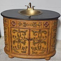 Front angle view of vintage vanity showing carved floral details, black countertop, brass sink and faucet, and front doors.