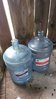 Two large empty reusable 5-gallon water bottles made of clear blue-tinted plastic, placed side by side on a wooden floor with rustic backdrop.