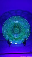 Full view of the vintage uranium glass plate illuminated under UV light, showing glowing green translucence and etched floral details.